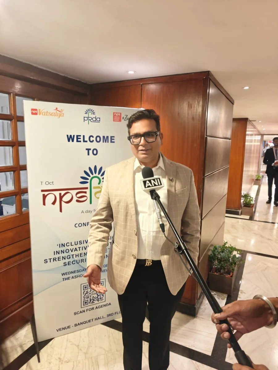 A man in a beige check blazer speaking to a reporter holding an ANI microphone, in front of the NPS Diwas conference welcome banner
