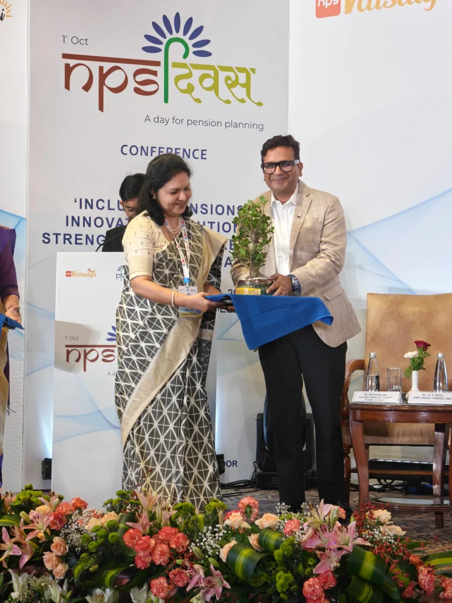A woman in a grey patterned saree presents a potted bonsai plant to a man in a beige blazer on stage at the NPS Diwas pension-planning conference
