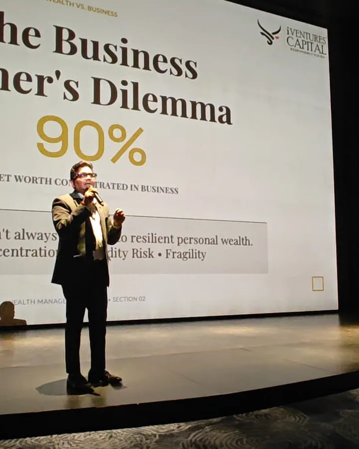 An iVentures speaker with a microphone on stage in front of a large slide titled 'The Business Owner's Dilemma', about business owners keeping most of their net worth tied up in their business